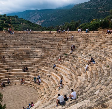 Tourists sitting on the stone steps of the ancient Greek theatre in Kas, Turkey, with mountain views.