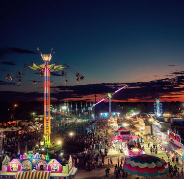 Aerial view of a vibrant night carnival with illuminated amusement park rides, games, and crowds at sunset.