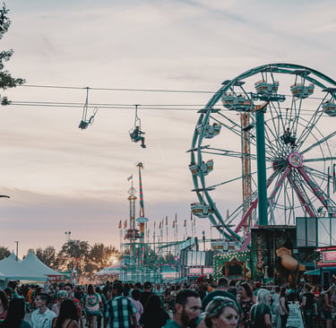 Crowds at a summer carnival with a Ferris wheel, chairlift ride, and ticket booths at sunset.