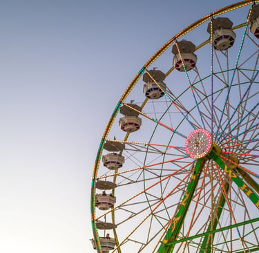 A large Ferris wheel with colorful LED lights spinning against a clear twilight sky at a local carnival.