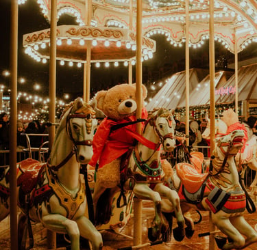 A large teddy bear in a red coat riding a vintage carousel horse at a festive outdoor carnival.
