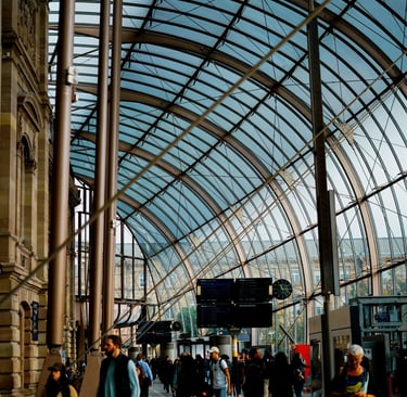 Commuters walk through a modern European train station under a large glass vaulted ceiling.