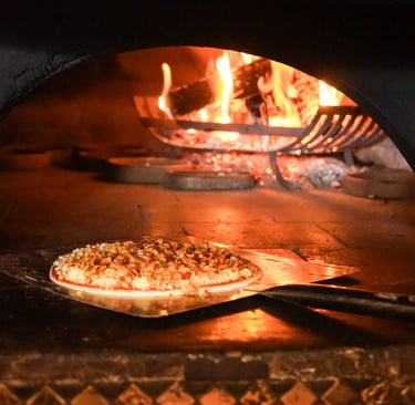 Authentic wood-fired pizza baking on a metal peel inside a stone oven with roaring orange flames.