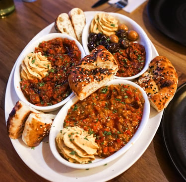 A Middle Eastern appetizer platter with hummus, shakshuka, olives, and warm pita bread on a wooden table.