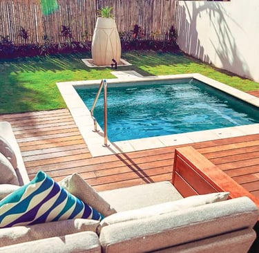 Compact square plunge pool with clear turquoise water and light stone coping around warm timber deck
