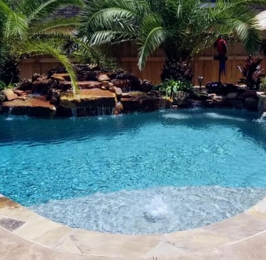 Curved tropical pool with rock waterfall, deep blue water and natural stone coping with pebble inter