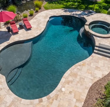 Organic-shaped pool with rich blue-green water and dark interior finish, bordered by beige stone til