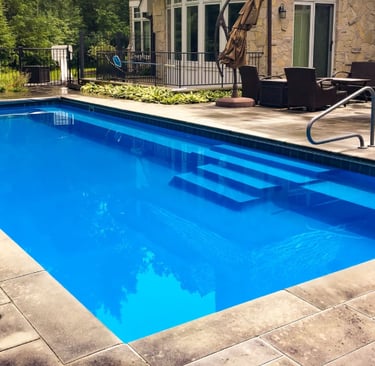 Modern rectangular pool with deep green-blue water and dark interior tiles, surrounded by natural st