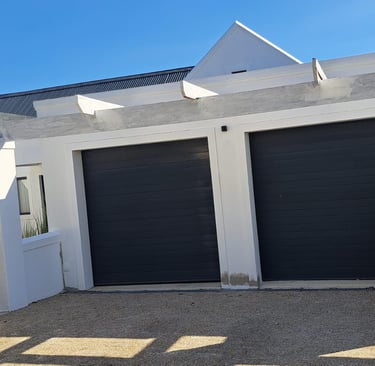 A professional painter applying a fresh coat of white paint to a modern house with two black garage doors.
