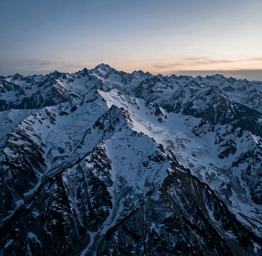 A high-altitude cinematic drone shot of a rugged mountain ridge at blue hour. The lighting transitions from deep #1B263B shadows to soft #A0B0C0 highlights on the snow-capped peaks. The composition is wide and sweeping, capturing professional 4K clarity and a sense of epic scale.