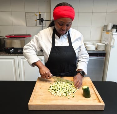 Chef Viviane in white chef jacket using a chef's knife to dice courgette on a bamboo chopping board on a dark kitchen surface