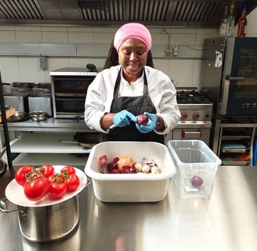 Chef Viviane in white chef jacket and black apron calmly peeling red onions beside fresh tomatoes on a stainless steel.