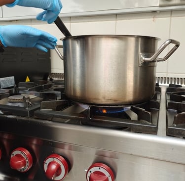 A chef's gloved hand stirring a large stainless steel stockpot over a blue gas flame on a professional kitchen range