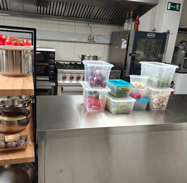 An organised professional kitchen station with pans on a wooden rack, fresh tomatoes and labelled prep containers.