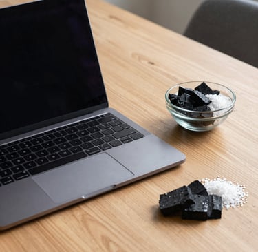 Laptop on a wooden desk next to a bowl of black charcoal and white sea salt.