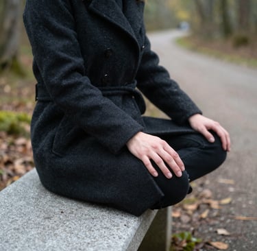 A person in a black wool coat sits in a cross-legged meditation pose on a stone park bench.