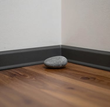 A smooth grey river stone sits on a walnut laminate floor in the corner of a room with white walls.