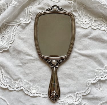 Vintage silver hand mirror with pearl accents on a white lace and linen background.