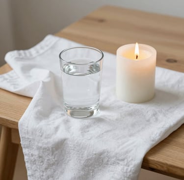 A glass of water and a lit white pillar candle on a white linen cloth over a wooden table.