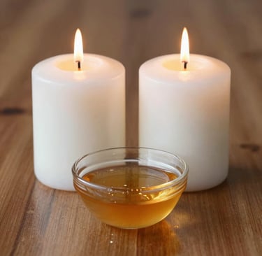 Two lit white pillar candles behind a small glass bowl of golden massage oil on a wooden table.