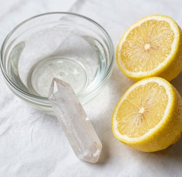 Clear quartz crystal point next to a glass bowl of water and two lemon halves on a white cloth.