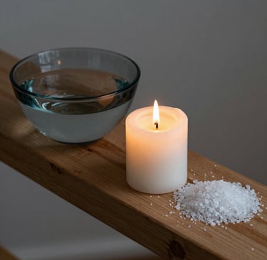 A lit white pillar candle beside a bowl of water and a pile of bath salt on a wooden spa tray.