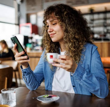 A curly-haired woman in a blue denim shirt using her smartphone while enjoying an espresso at a cafe.