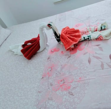 Two decorative swan towels folded into a heart shape on a pink floral bedsheet in a hotel room.