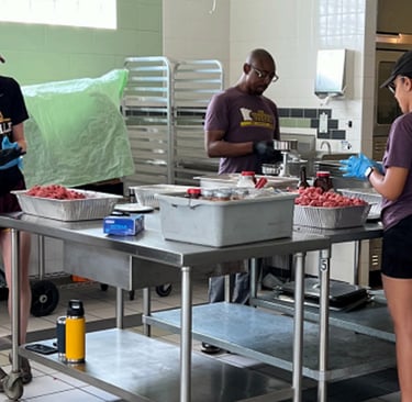 Volunteers in purple shirts preparing food together in a professional commercial kitchen setting.