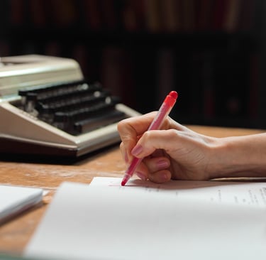 A person using a red pen to edit a manuscript next to a vintage typewriter in a library.