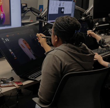 a creative student sitting at a desk with two monitors and a laptop