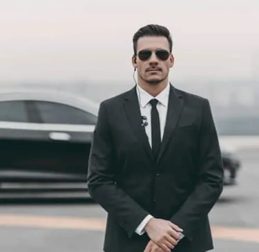 En livvakt står i kostym och solglasögon framför en svart bil.