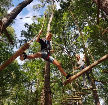 Two children wearing safety harnesses navigate a high ropes obstacle course in a forest park.