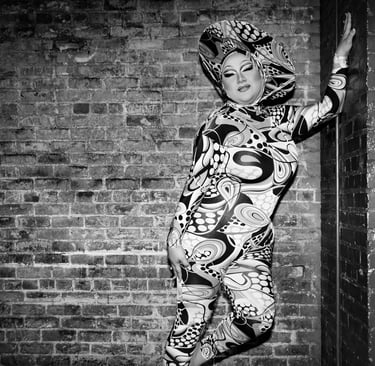 black & white drag queen posed fashionably in front of brick wall on stage in a patterned bodysuit