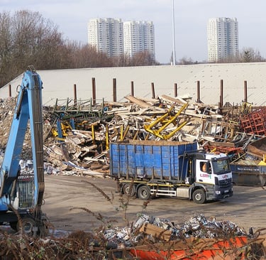 A scrap metal recycling yard with piles of mixed metal, wood, and industrial debris. A blue hydrauli