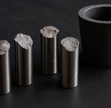 Four fractured metal fatigue test specimens beside a graphite crucible.