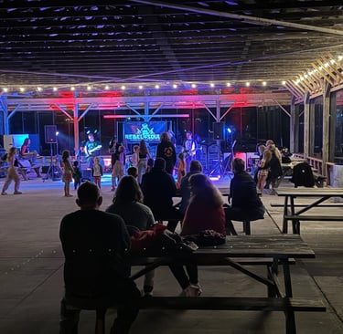 Live band performing for a crowd under a lit outdoor pavilion with picnic table seating at night.