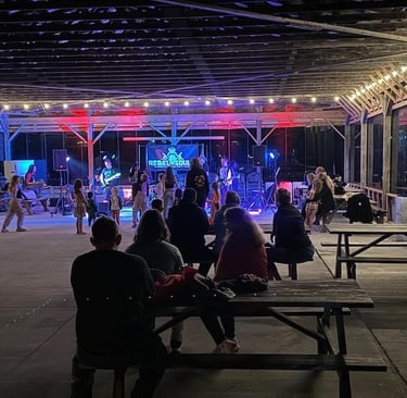 Live band playing for an audience under a lit pavilion with picnic tables at an outdoor concert event.