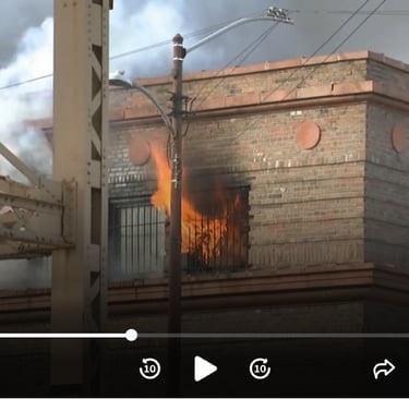 Flames and smoke pour out of a brick building window during a structure fire with active firefighting efforts.