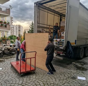 Antalya'da bir kamyonun arkasından büyük bir ahşap paneli indiren ve taşıma arabasına yükleyen işçiler.
