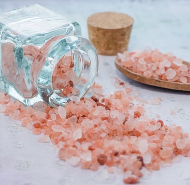 Pink Himalayan salt spilled from a glass jar onto a surface with a wooden spoon nearby.