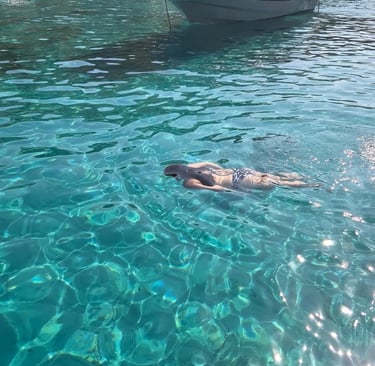 A woman swimming in crystal clear turquoise water near a white motorboat in a Mediterranean cove.