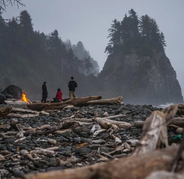 a group of people standing around a campfire in the PNW among big rocks and the ocean