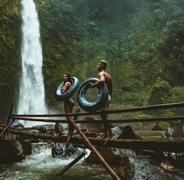 a woman and a man standing on a bridge by a waterfall holding inner tubes