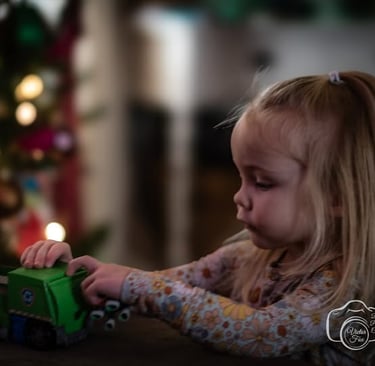 A young blonde girl playing with a green toy truck near a blurred Christmas tree.