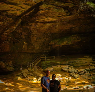 A couple and their black dog stand before a large cave entrance to a small waterfall, Ash Cave Logan