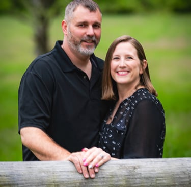 A smiling couple posing for an outdoor portrait by a rustic wooden fence in a lush green field.