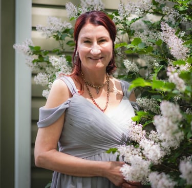 A woman in a grey formal gown smiling outdoors next to blooming white lilac flowers.