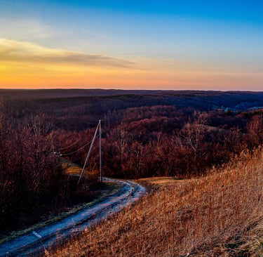 Winding dirt road through a hilly autumn forest landscape at sunset with vibrant orange and blue sky.