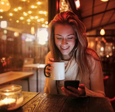 Smiling woman holding a coffee mug while looking at her smartphone in a cozy, warmly lit café at night.